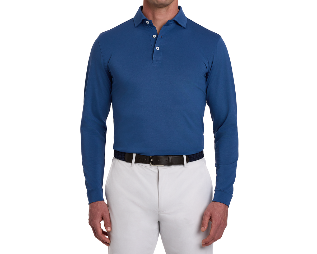 The Abbott Shirt: Andover, a long-sleeve polo in blue, worn with gray trousers. Features a spread collar, contrast piping, and breathable performance fabric.