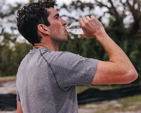 Young man outdoors in profile wearing The Pickett Performance T-Shirt, Heathered Gray, drinking from a water bottle, showcasing breathable, moisture-wicking fabric.