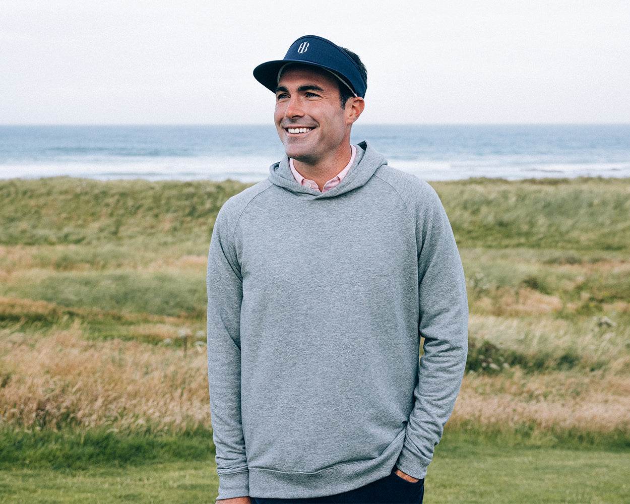 Man in a seaside setting wearing The Lawson Hoodie: Heathered Gray, a light-gray pullover, ideal for cool days, without drawstrings, over a collared shirt.