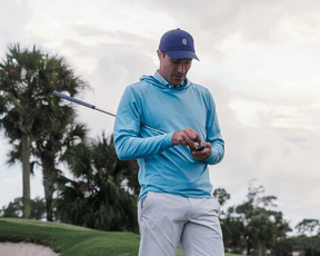 Man wearing The Jackson Hoodie: Harbor, a lightweight pullover, on a golf course, holding a smartphone with a golf club nearby.