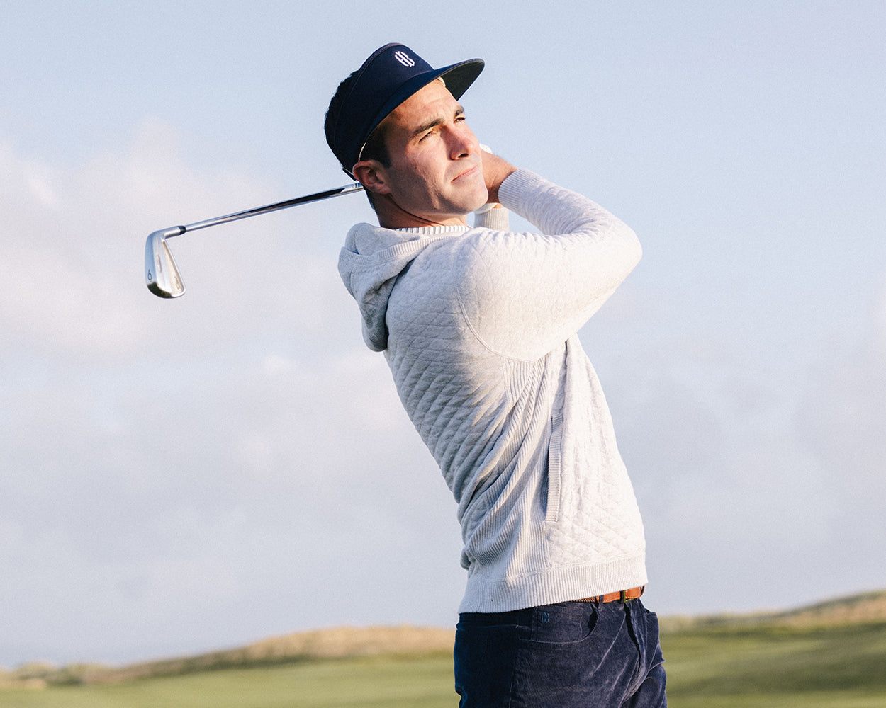 Man on golf course wearing The Wallace Hoodie Sweater: Heathered Gray, mid-swing with golf iron, showcasing its quilted design and streamlined fit.