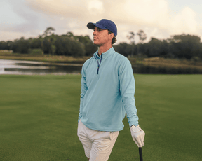 Man on golf course wearing The Stuart Quarter-Zip: Harbor, holding a golf club, showcasing its lightweight, textured rib fabric and superior softness.