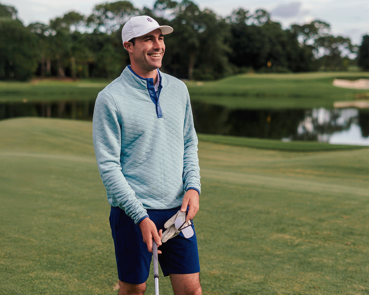 Man on golf course wearing The Sullivan Quarter-Snap: Heathered Harbor, a light-blue quilted pullover with diamond insulation, holding a golf glove and putter.