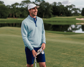 Man on golf course wearing The Sullivan Quarter-Snap: Heathered Harbor, a light-blue quilted pullover with diamond insulation, holding a golf glove and putter.