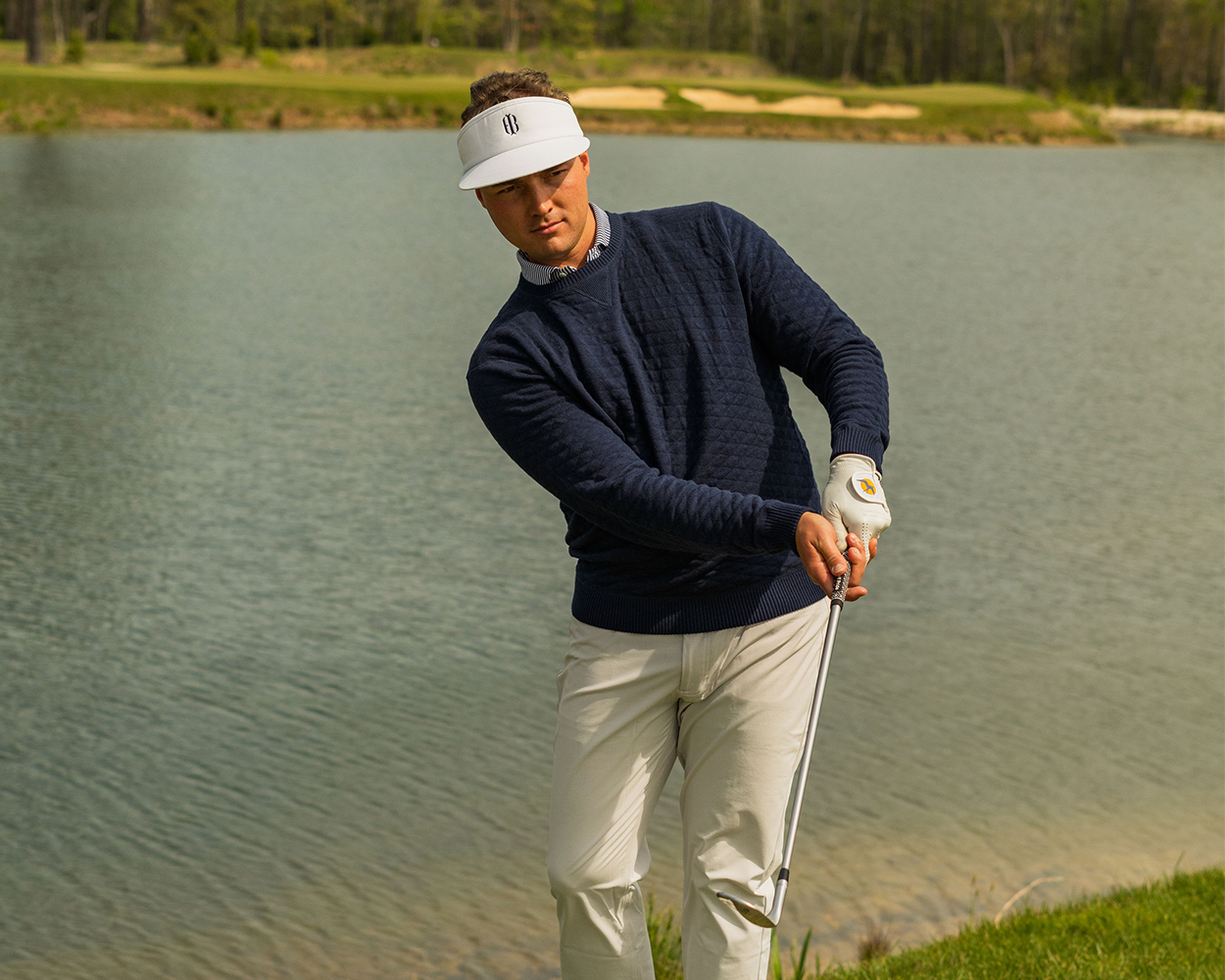 Man chips shot at pond edge in golf course, wearing The Ward Crewneck Sweater: Heathered Navy, showcasing its diamond jacquard knit pattern and versatile style.