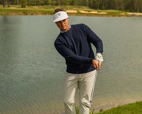 Man chips shot at pond edge in golf course, wearing The Ward Crewneck Sweater: Heathered Navy, showcasing its diamond jacquard knit pattern and versatile style.