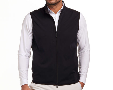 Man wearing The Ventura Vest: Black, a sleeveless zip-front vest, over a white shirt. Features perforated fabric and zippered pockets for functionality.