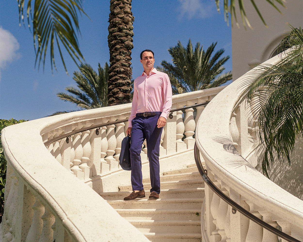Man wearing The Goodrich Shirt: Belmont on a white stone spiral staircase, holding a jacket, with palm trees in the background, showcasing its stylish, versatile design.