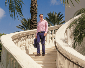Man wearing The Goodrich Shirt: Belmont on a white stone spiral staircase, holding a jacket, with palm trees in the background, showcasing its stylish, versatile design.