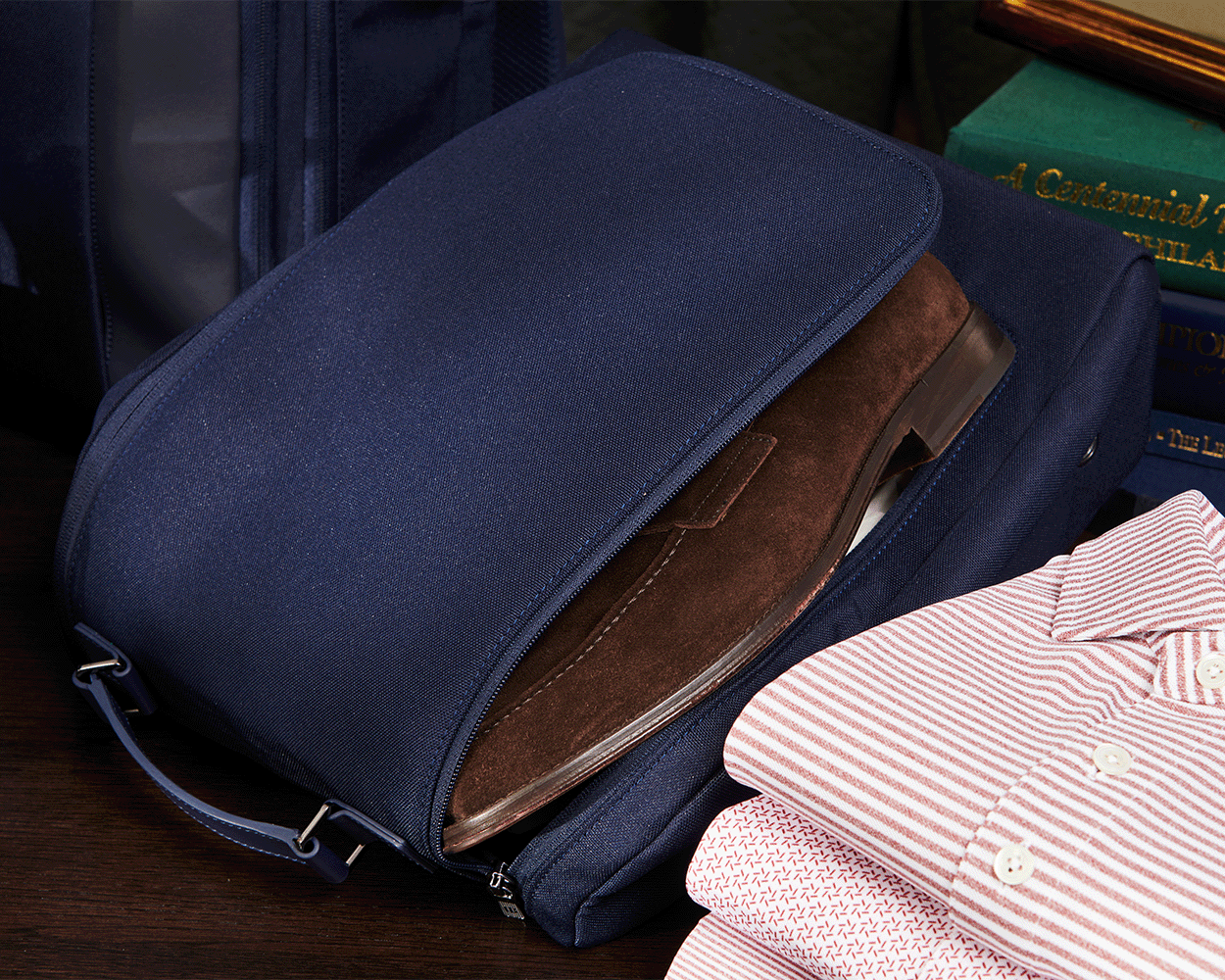 The Rutledge Shoe Bag: Navy Ballistic, open with a brown suede shoe visible, beside folded shirts on a wooden surface, showcasing its durable design and leather trim.
