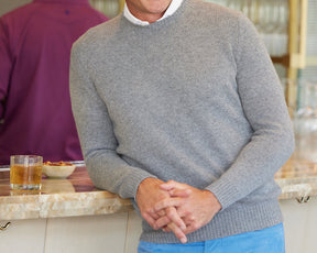 Man in The Berwick Crewneck Sweater: Heathered Gray, leaning on counter with drink and nuts; showcases classic crewneck style and plush cashmere texture.