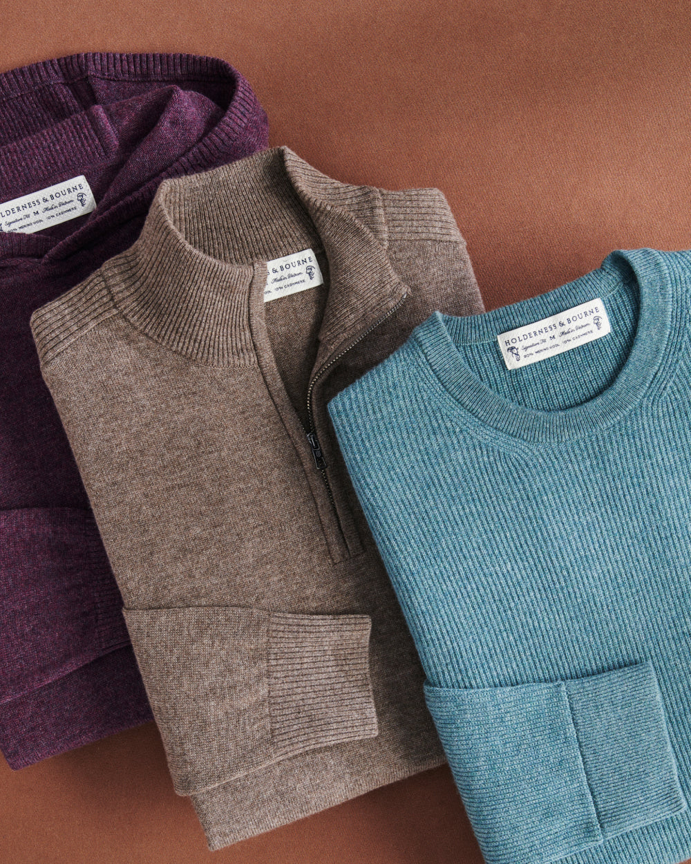 Folded sweaters in plum, taupe quarter-zip and teal ribbed crewneck on a rust-colored background.