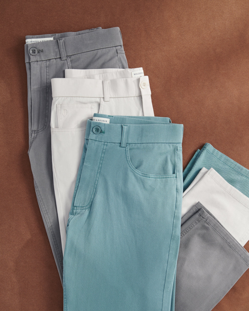 Folded five-pocket pants in gray, off-white and teal on a rust-colored background.