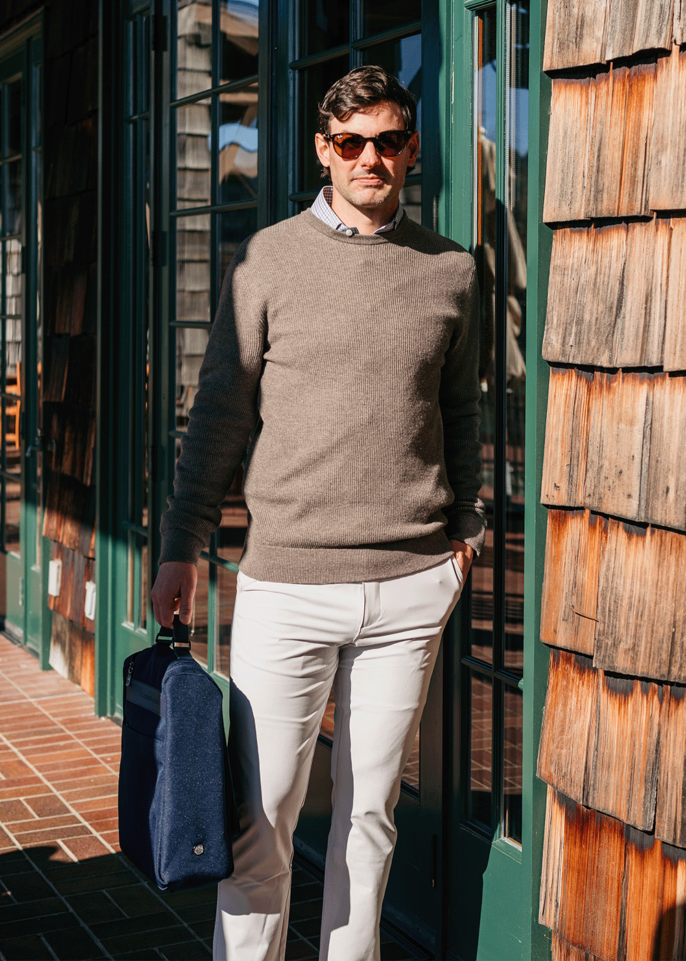 Man in sunglasses wearing a taupe crewneck sweater and white chinos, carrying a navy bag outside a green-framed shingled building.