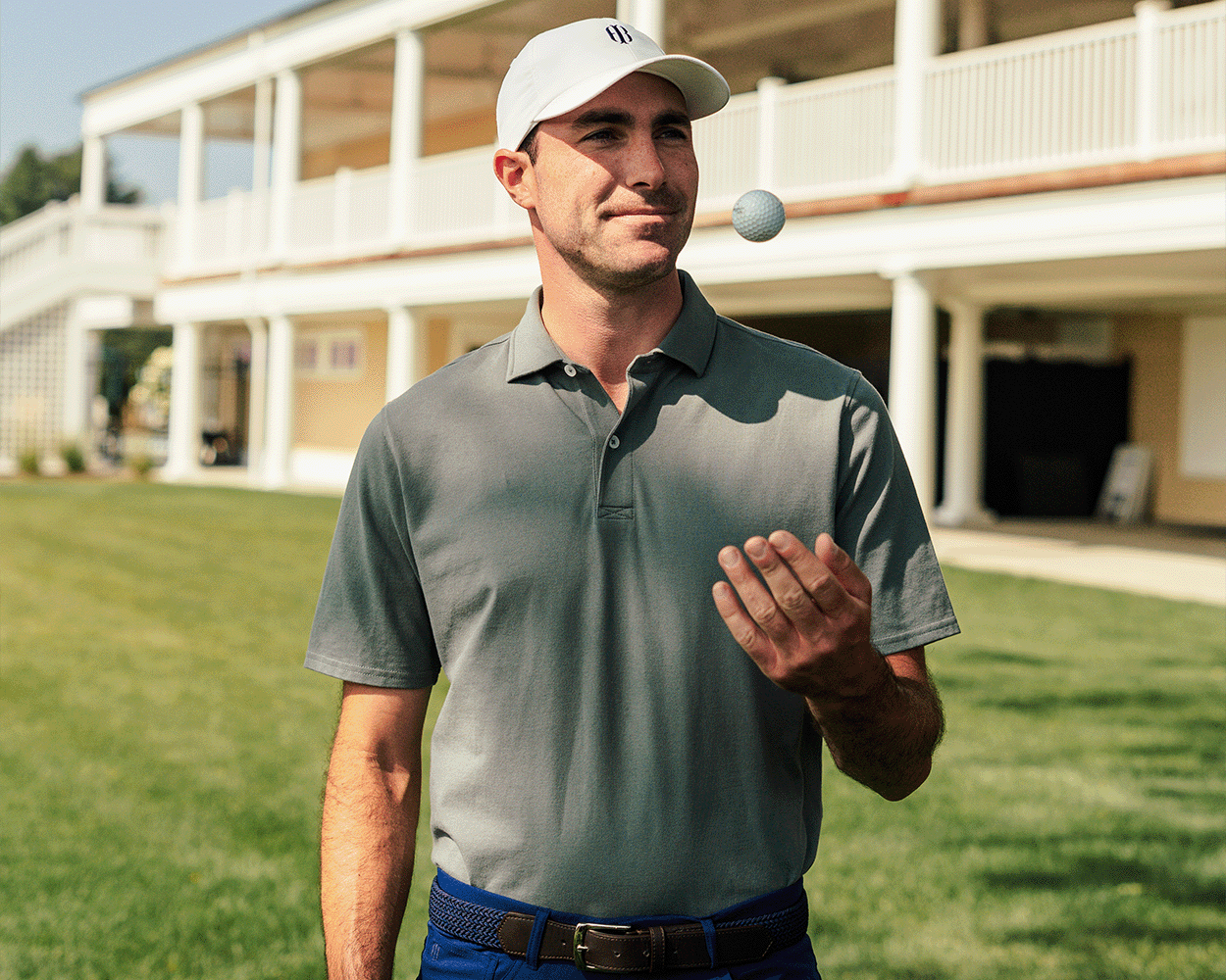 Man in golf attire wearing The Neville Shirt: Ivy, a soft, durable polo, tosses a ball in front of a clubhouse setting.