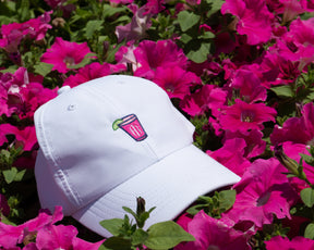 The Lightweight Performance Hat: White with Transfusion Icon, displayed among pink flowers, features a pink drink embroidery, adjustable fit, and UPF 50+ protection.