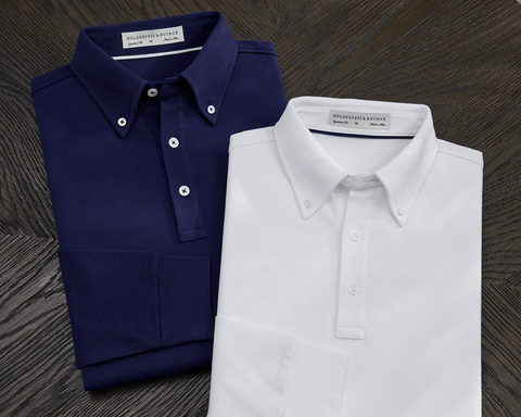 The Burton Shirt: White, a long-sleeve polo with a button-down collar, three buttons, and stretch ribbed cuffs, displayed alongside its navy counterpart on a wooden surface.