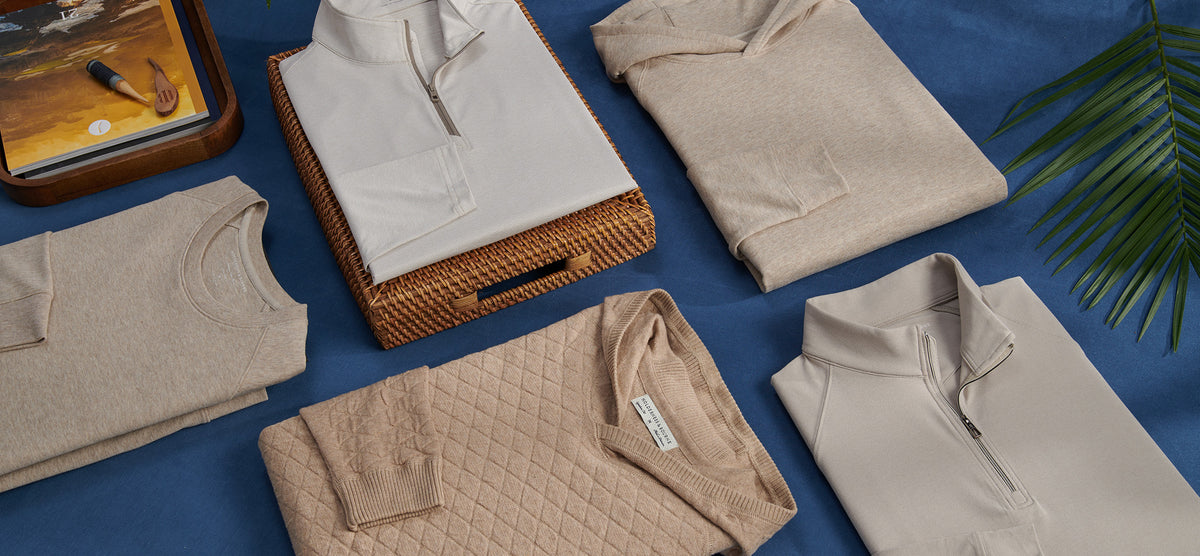 Flat lay of folded neutral sweaters, quarter-zips and a quilted pullover on a blue cloth with a palm frond and a book.