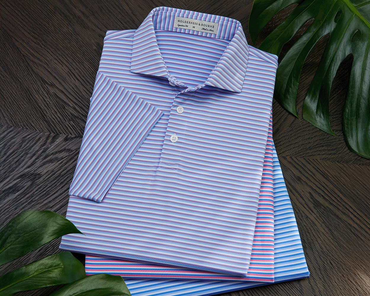Stack of The Fleming Shirt in fine horizontal stripes, showcasing its structured spread collar and signature fit, surrounded by decorative green leaves on a dark wood surface.