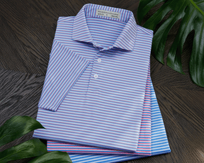 Stack of The Fleming Shirt in fine horizontal stripes, showcasing its structured spread collar and signature fit, surrounded by decorative green leaves on a dark wood surface.