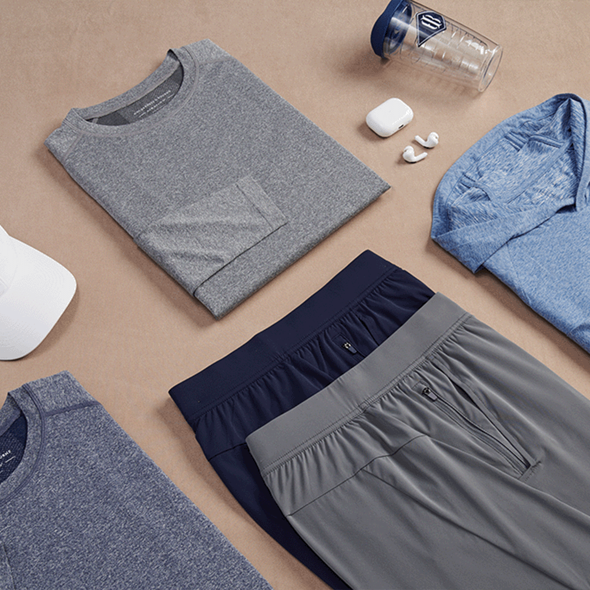 Folded gray shirt, navy shorts, gray pants, blue shirt, white earbuds, and a clear water bottle on a beige surface.