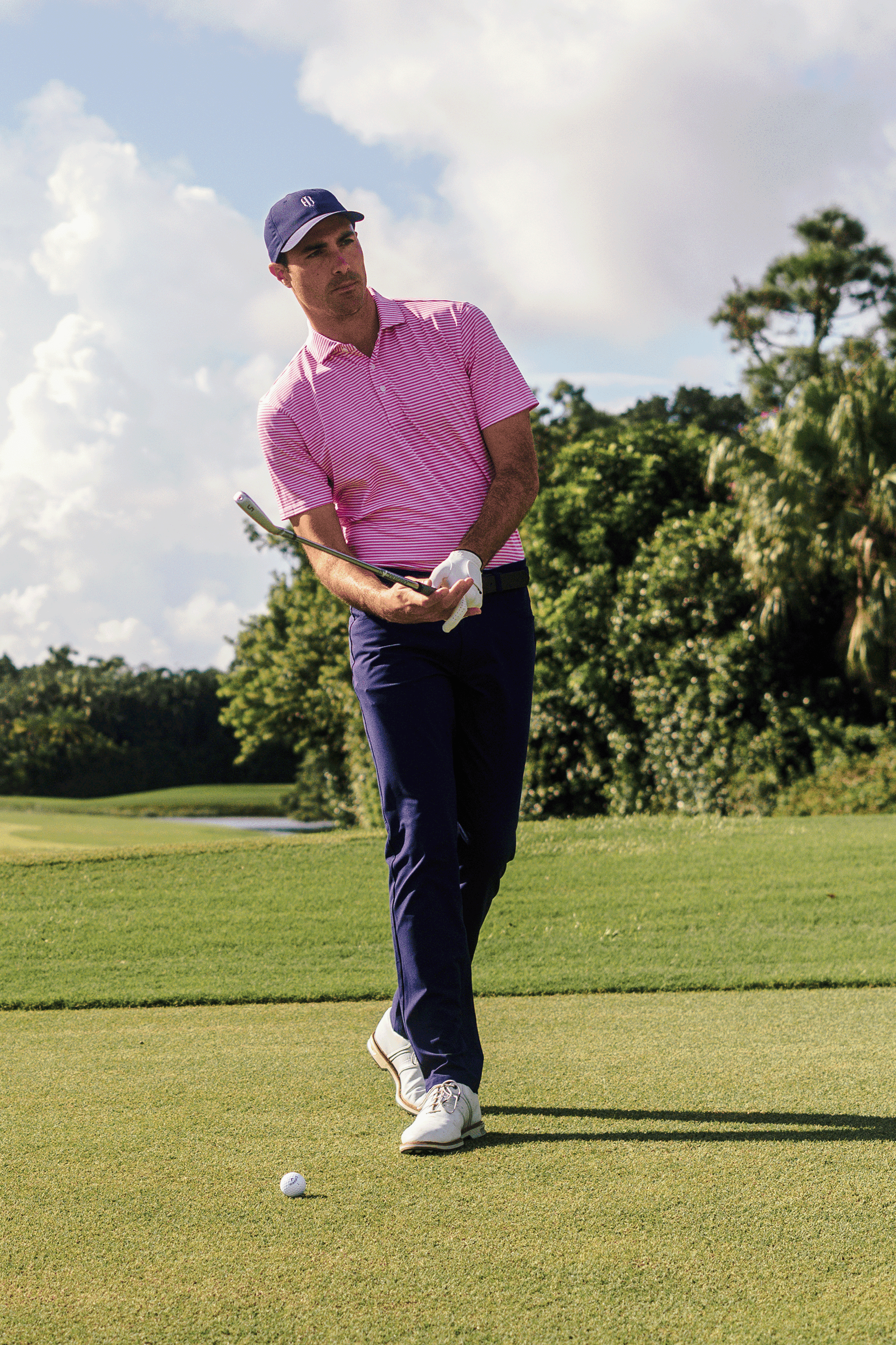 Man in pink shirt and blue pants playing golf on a green course with trees in the background