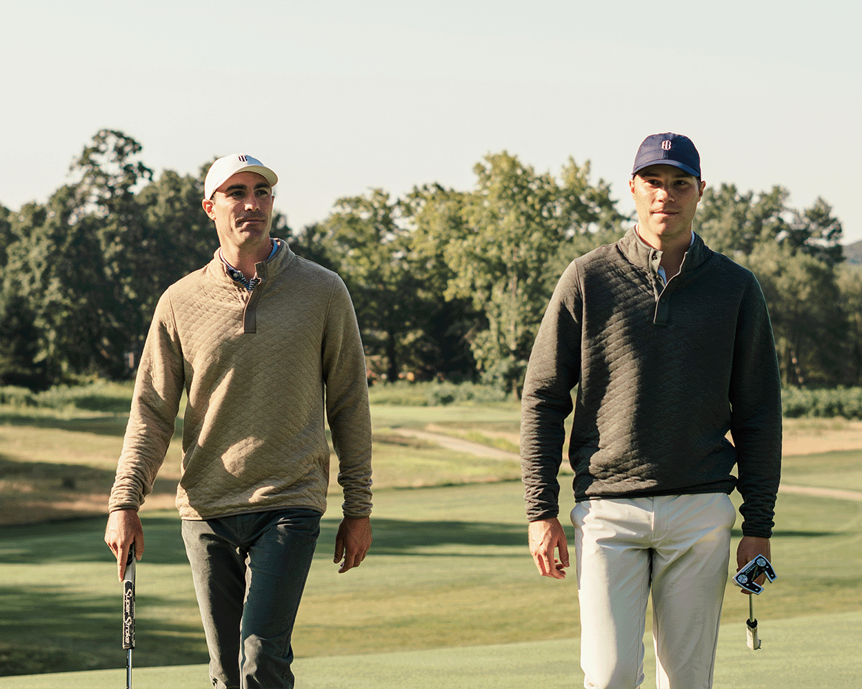 Two men on a golf course wear The Sullivan Quarter-Snap: Heathered Dune pullovers, showcasing its quilted texture and classic style while carrying putters.