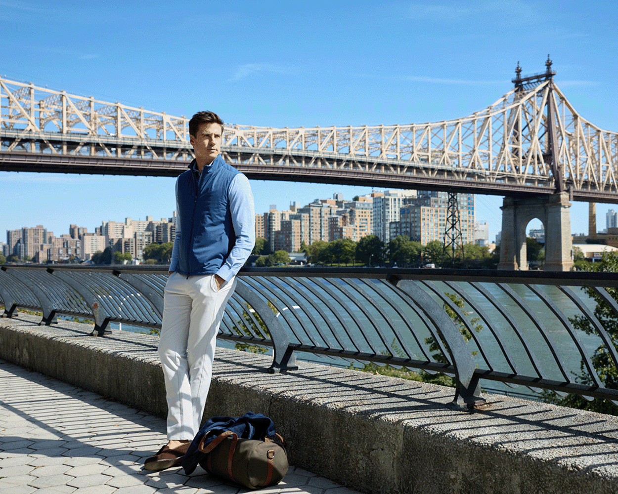 A young man wears The Swenson Vest: Andover, a quilted vest ideal for warmth and breathability, leaning on a riverside railing with a duffel bag nearby.