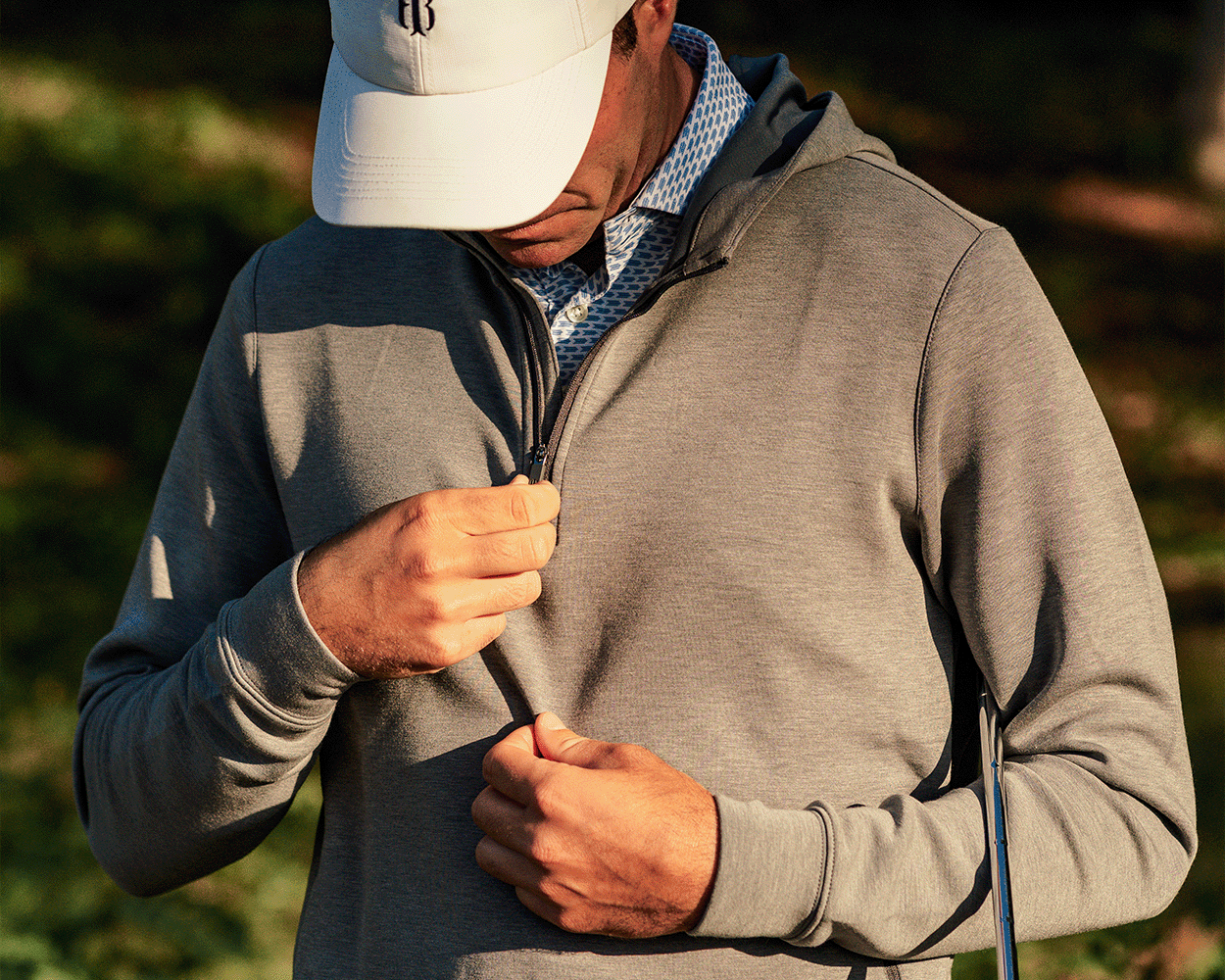Man outdoors wearing The Tilley Quarter-Zip Hoodie, pulling up the zipper, with a golf club under his arm, suggesting readiness for a golf game.