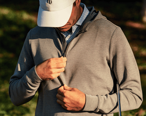 Man outdoors wearing The Tilley Quarter-Zip Hoodie, pulling up the zipper, with a golf club under his arm, suggesting readiness for a golf game.