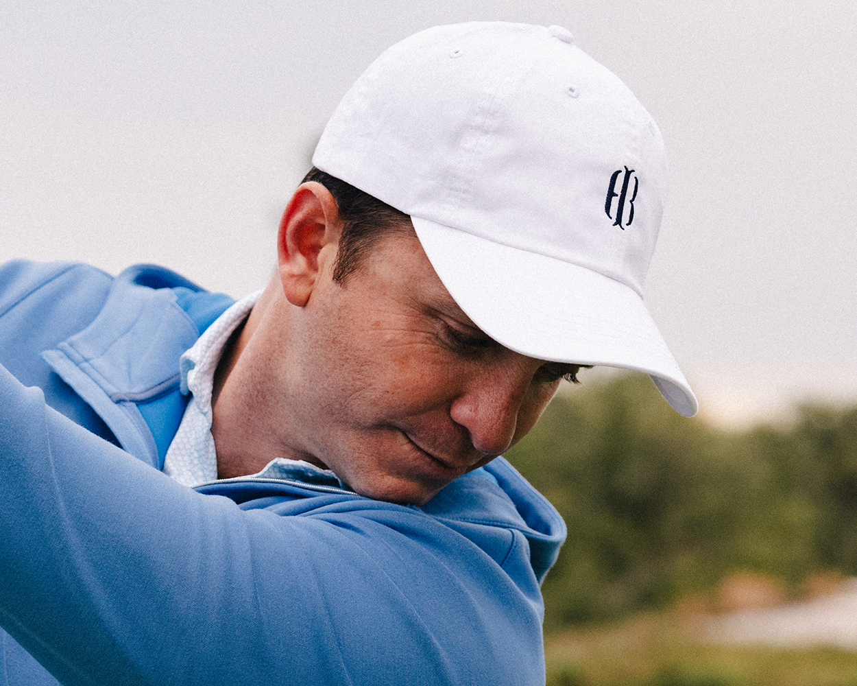 Man mid-swing wearing The Lightweight Cotton Hat: White with HB Icon, featuring a low profile, embroidered FB, and adjustable closure for comfort.