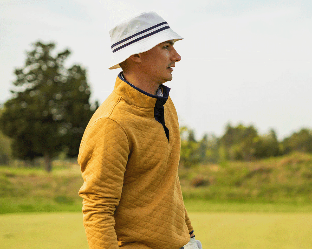 Young man on a golf course wearing The Lightweight Bucket Hat: White with HB Icon, featuring striped grosgrain ribbon and a high crown.